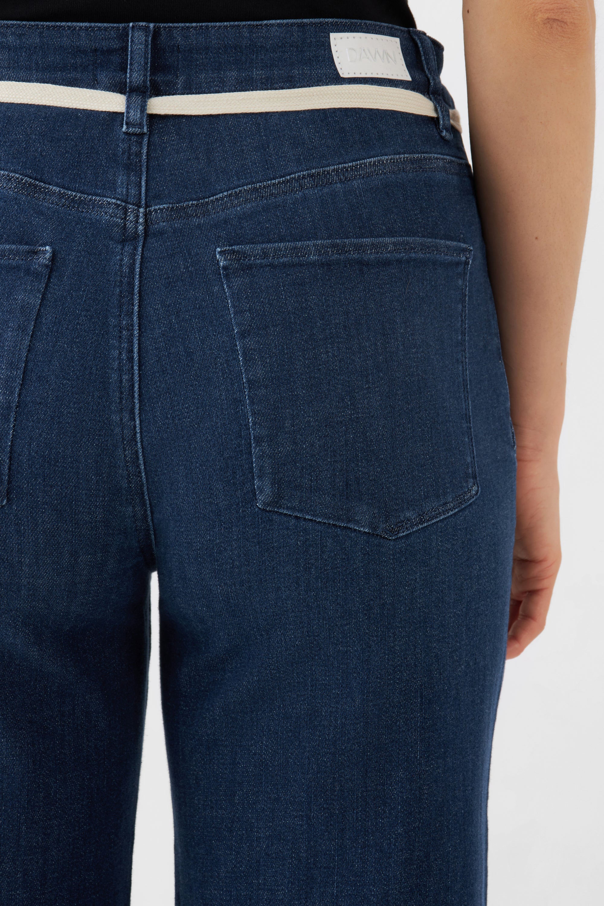 Rückansicht einer dunkelblauen DEW Flared-Jeans mit hoher Taille, zwei Gesäßtaschen, hellem Bundstreifen und DAWN-Label.