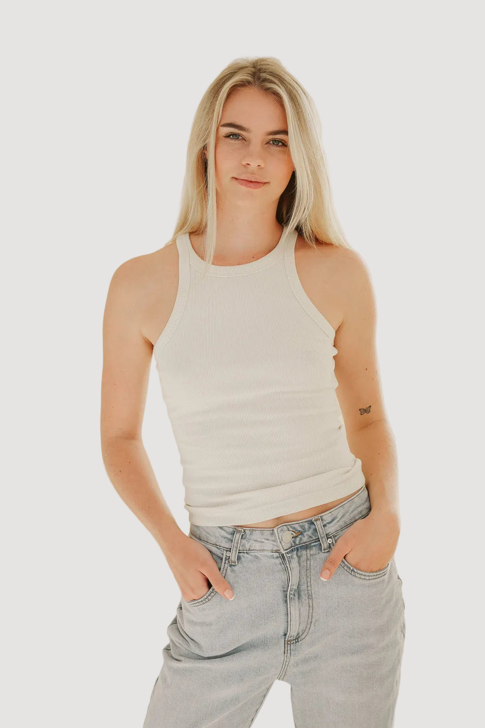 A woman wearing a white tank top and light blue denim jeans standing against a plain background.