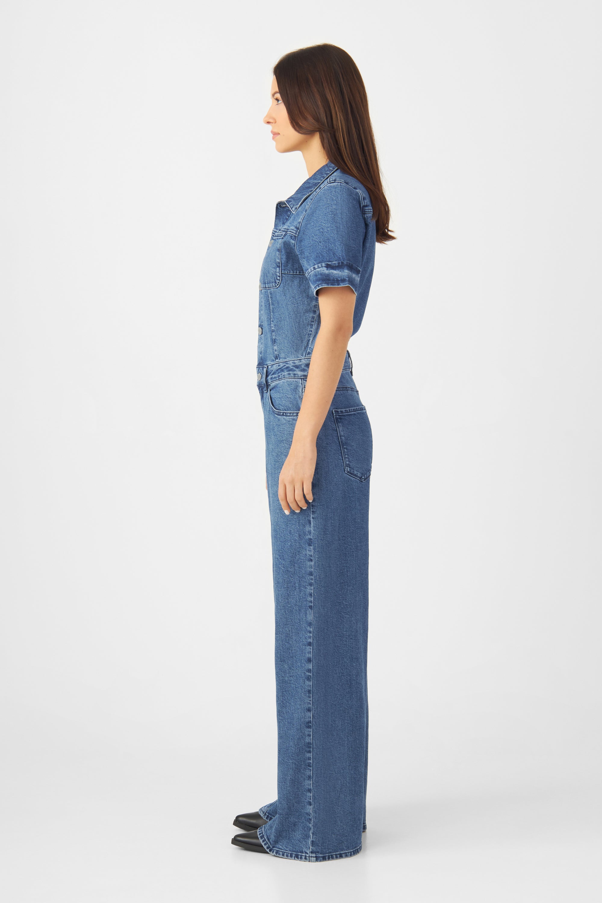 Seitenprofil eines Models in einem mittleren Blau Denim-Overall mit kurzen Ärmeln, taillierter Passform und weiterem Bein.