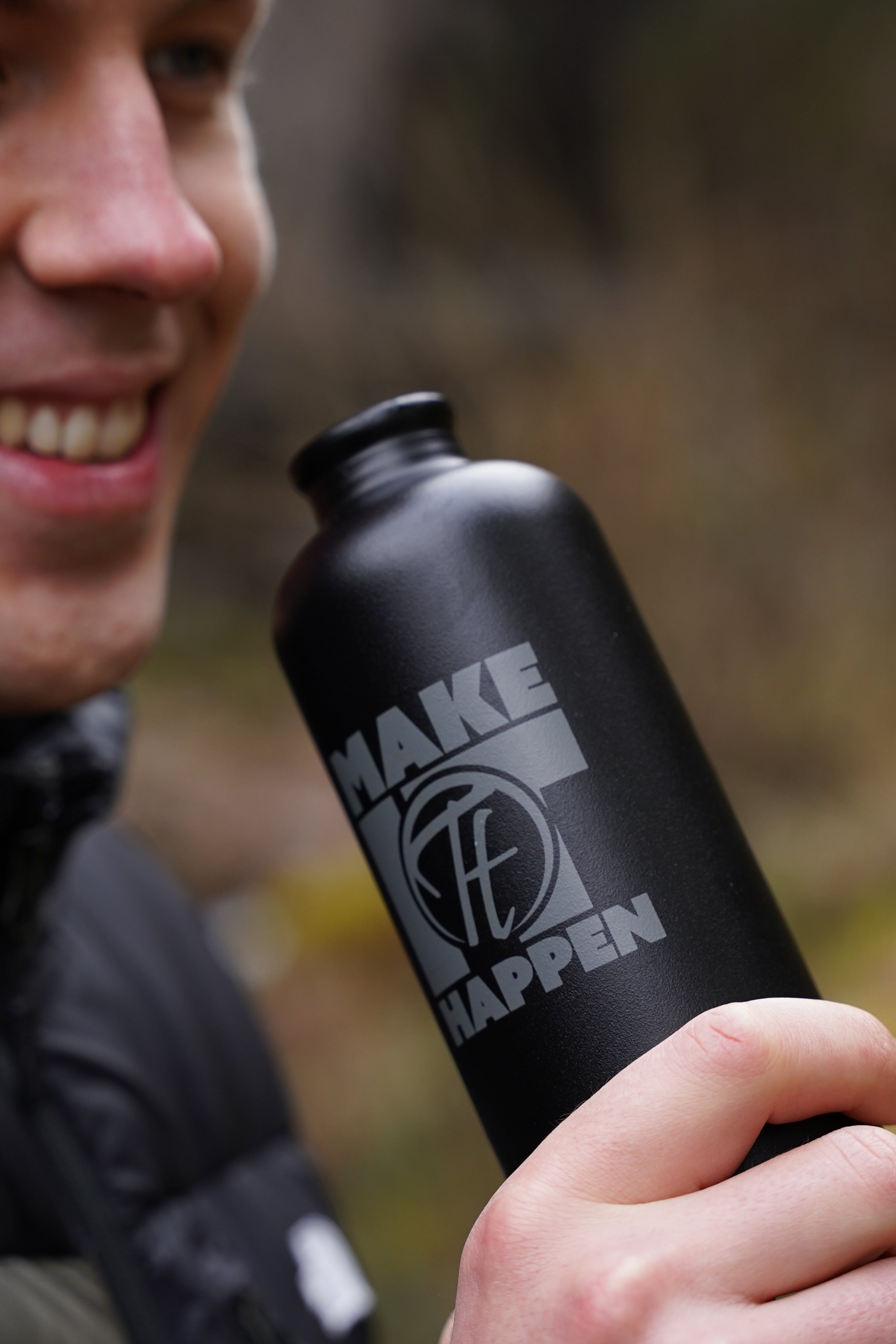 Schwarze Aluminium Trinkflasche aus 100% recycelten Materialien mit einem edlen "Make it happen" Print, draussen fotografiert. Ein Mann hält die Flasche in der Hand und lächelt; er ist kurz davor, einen Schluck zu trinken