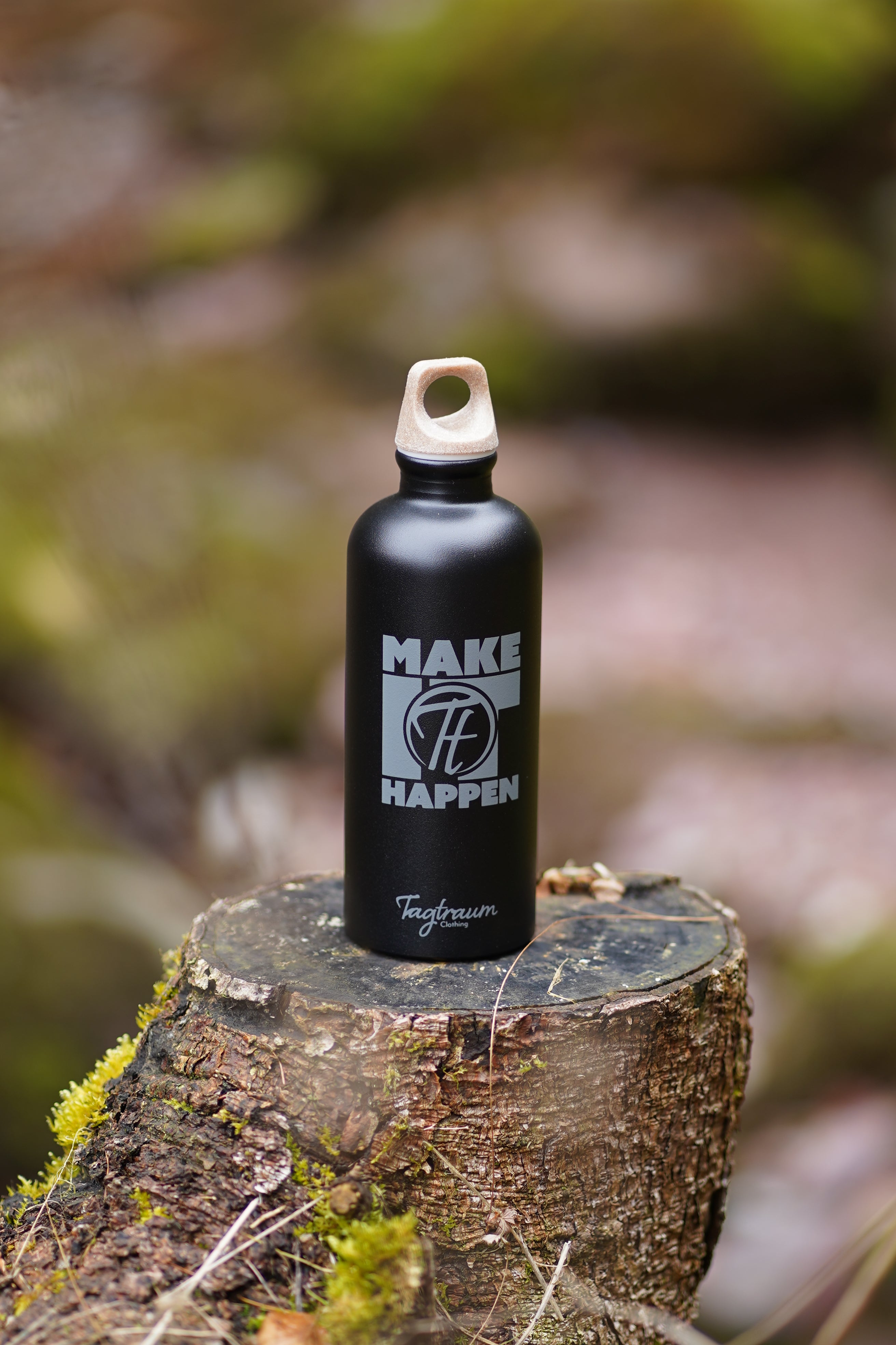 Schwarze Aluminium Trinkflasche aus 100% recycelten Materialien mit einem edlen "Make it happen" Print und einem Verschluss aus Cellulose, in Szene gesetzt draussen auf einem abgesägten Baumstrunk