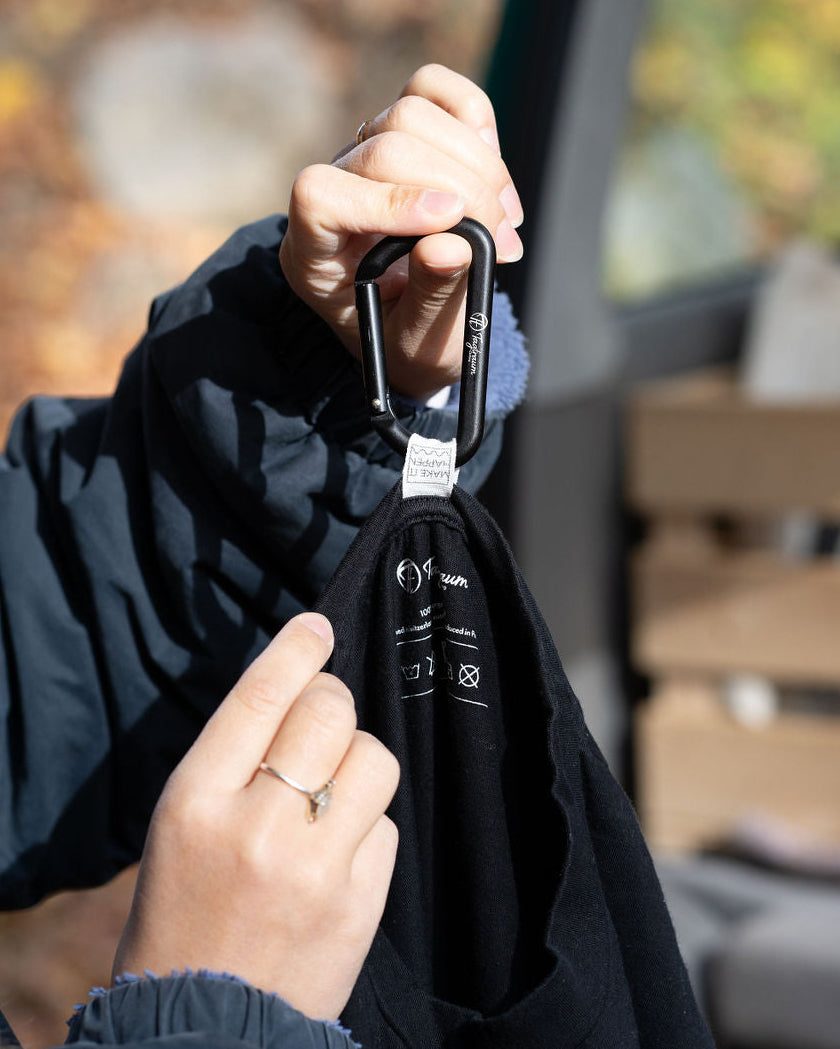 Ein kleiner Karabiner in Schwarz, an dem ein Tagtraum T-Shirt hängt, welches eine Make-It-Happen-Lasche am Nacken hat, um den Karabiner einzufädeln. So lassen sich alle Produkte von Tagtraum Clothing ganz einfach und überall aufhängen. 